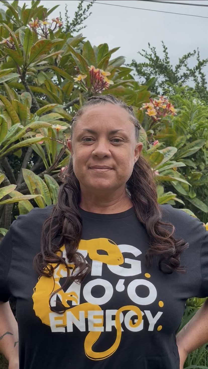 Kiʻi Kahoʻohanohano standing among plumeria, wearing a Big Moʻo Energy shirt.